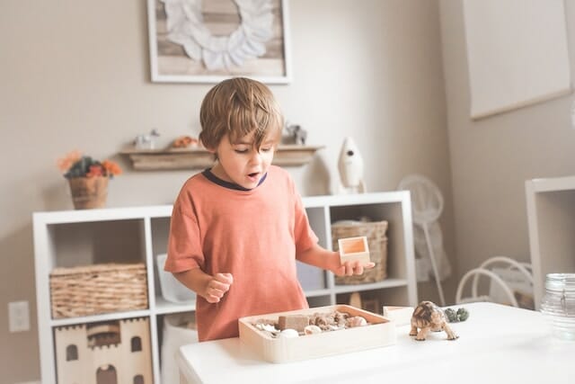 L'image montre un enfant, assis à une petite table, qui manipule un cube de bois Montessori. L'enfant est concentré sur la tâche et semble intéressé par l'objet. Il utilise ses mains pour explorer les différentes formes et textures du cube, et semble découvrir de nouvelles choses à chaque fois qu'il le manipule. À côté du cube se trouve un autre objet Montessori, un plateau avec des perles de couleurs différentes. L'enfant semble également attiré par ce plateau et pourrait bientôt s'intéresser à cette nouvelle activité. Les objets Montessori sont conçus pour aider les enfants à développer leur motricité fine, leur créativité et leur curiosité, et cet enfant semble déjà en profiter pleinement.